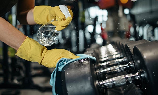 person cleaning gym equipment with a cloth and spray bottle